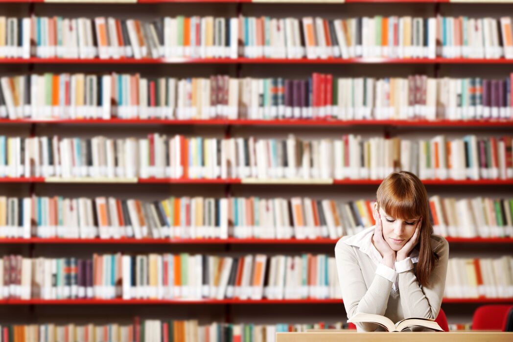 Das Bild zeigt eine junge Frau, die in der Bibliothek ein Buch liest.
